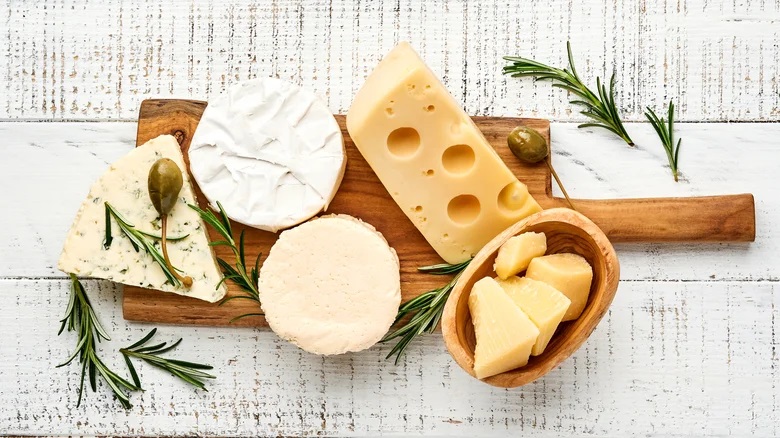  Slices of different cheeses on a wooden cedar board with olive leaves next to it